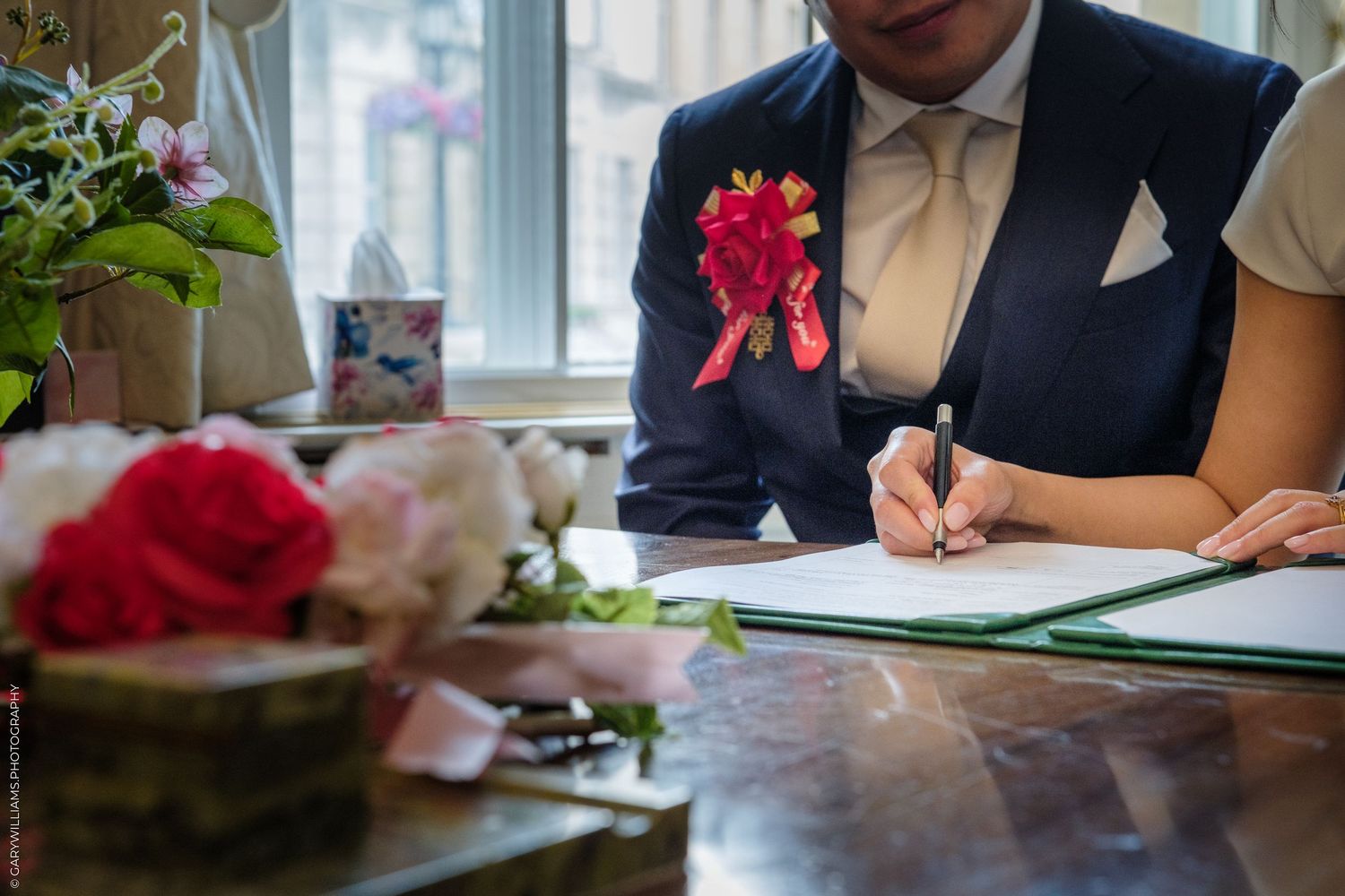 Groom in navy suit with floral boutonniere signing wedding documents at a table decorated with red and white roses.