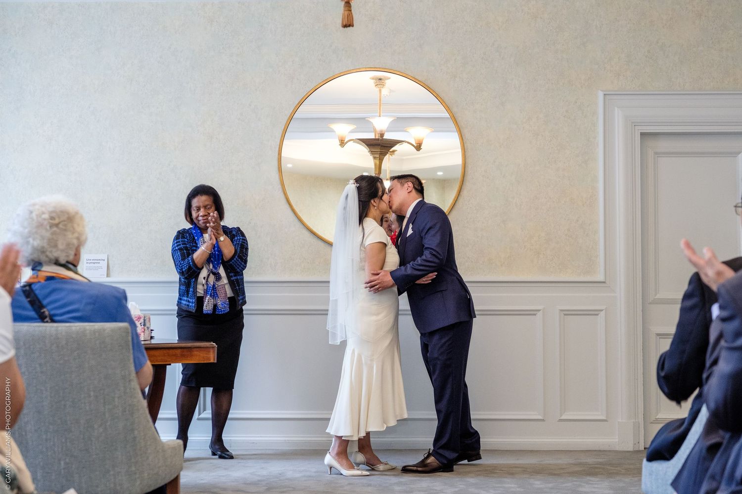 Couple shares first kiss at wedding ceremony while guests applaud in elegant white paneled room.