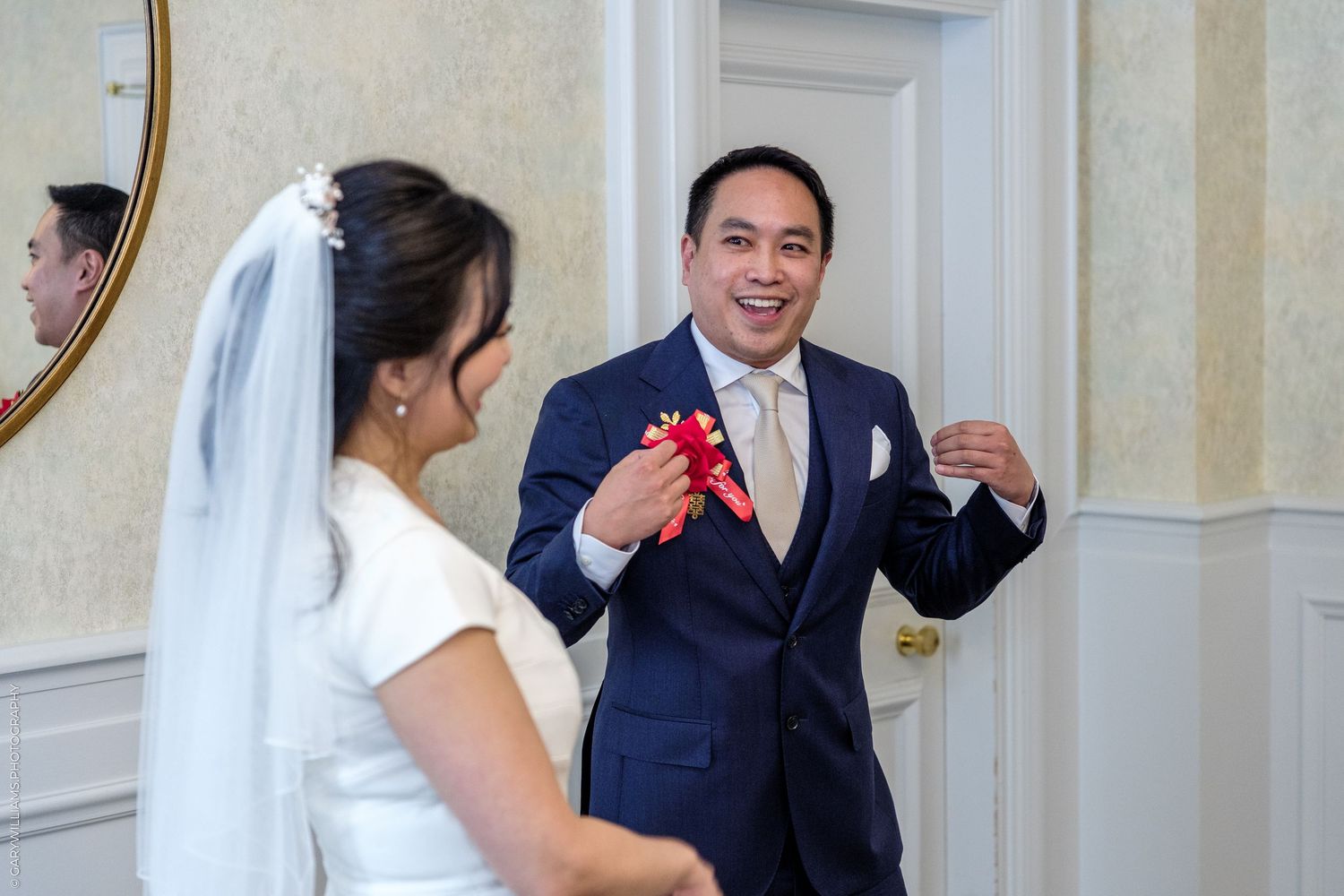 Groom in navy suit presenting a red gift to bride in white dress and veil during wedding ceremony.