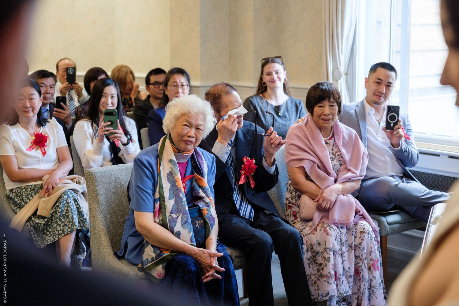 Elderly guests and attendees seated at a formal indoor event, some holding phones to capture the occasion.
