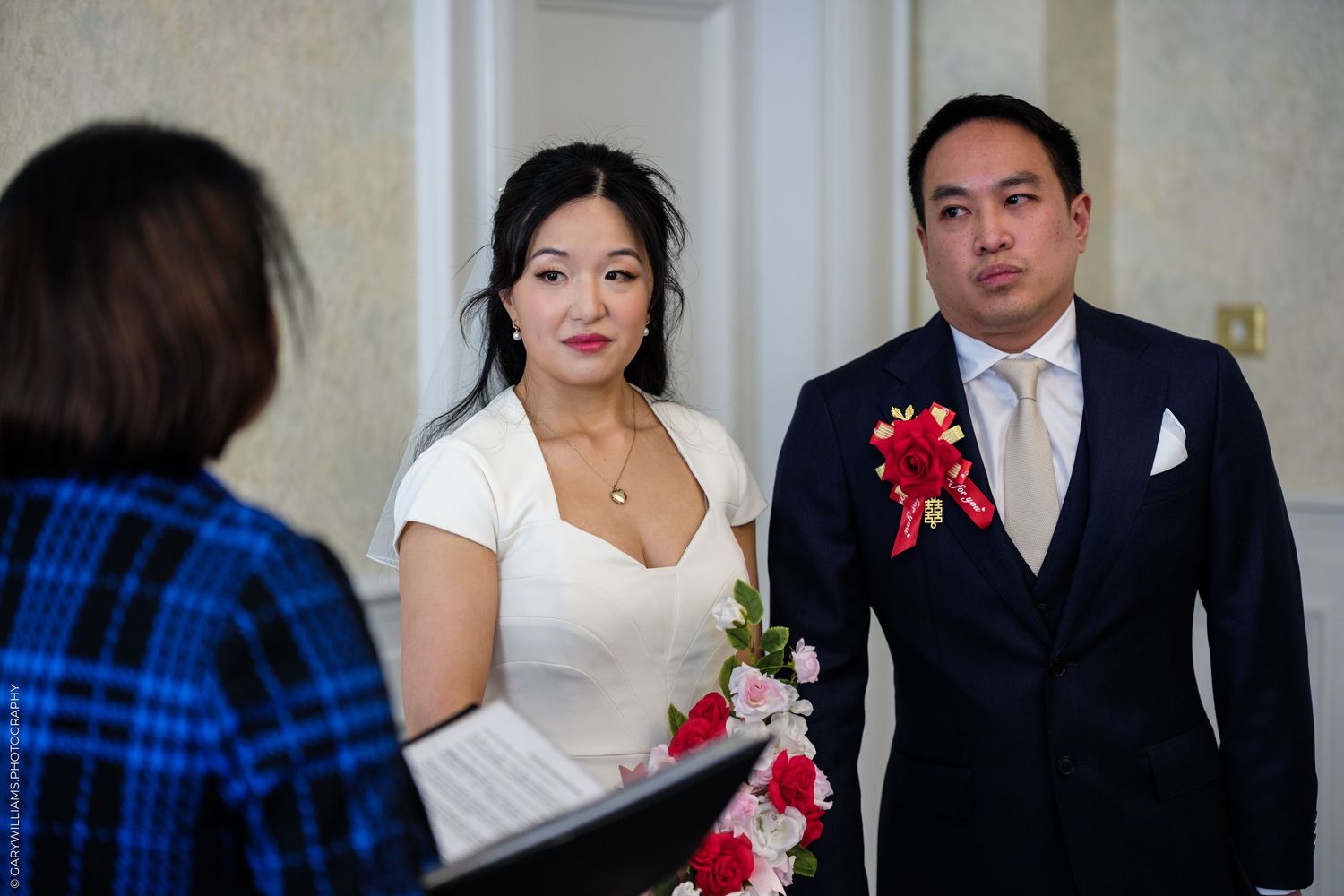 Asian couple stands before officiant during intimate wedding ceremony, groom in suit with red boutonniere.