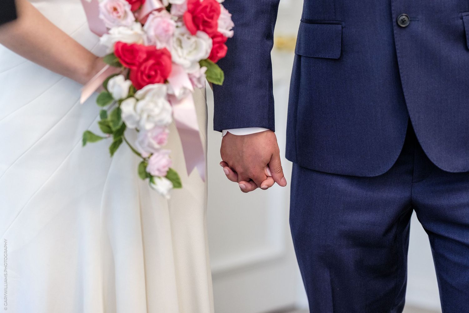 Bride and groom holding hands at wedding ceremony, bride holding pink and red rose bouquet, groom in navy blue suit.