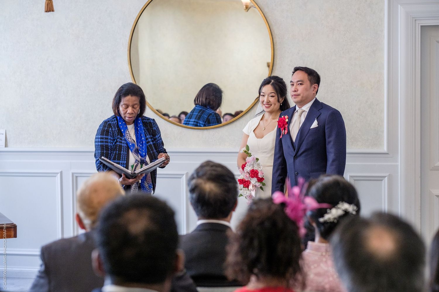 Officiant reading vows to Asian couple during small wedding ceremony with seated guests.