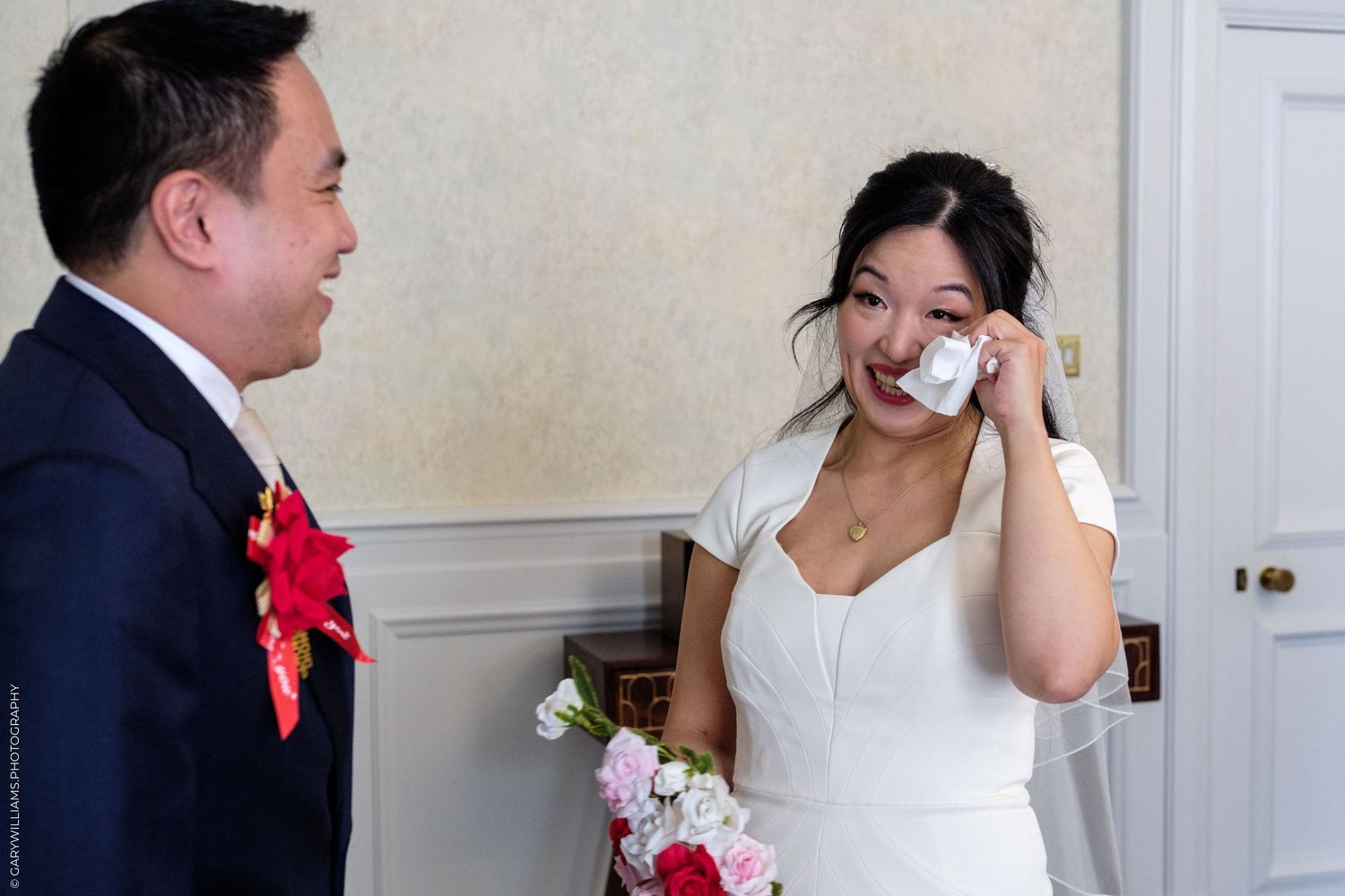 Emotional bride wiping tears while groom looks on during wedding day.