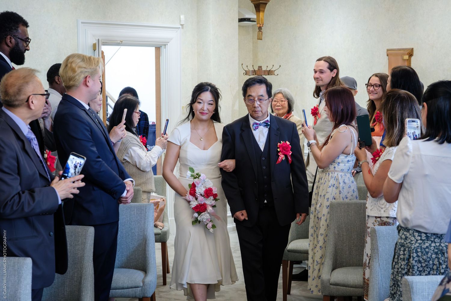 Asian couple walks down wedding aisle in elegant venue, bride holding red bouquet, guests applauding and taking photos.