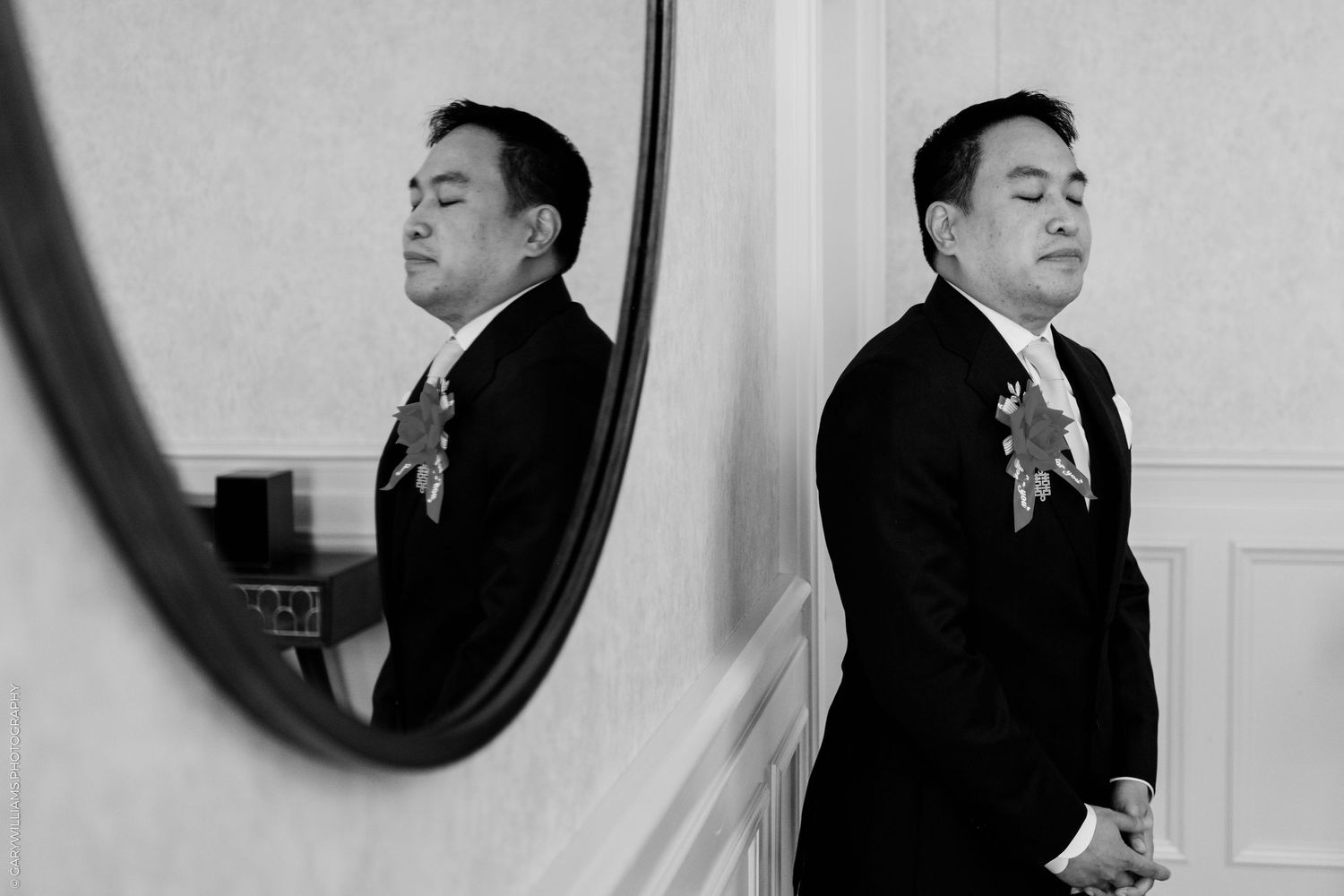 Black and white photo of a groom in a suit with boutonniere reflected in a round mirror before wedding ceremony.