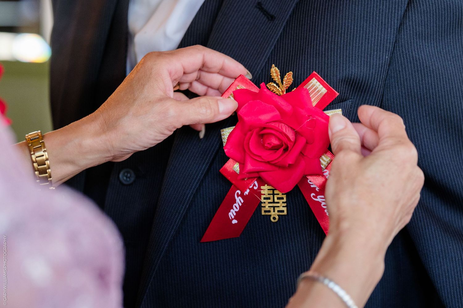 Hands pinning a red rose boutonniere with gold leaves and red ribbon onto a man's dark suit jacket at a wedding.
