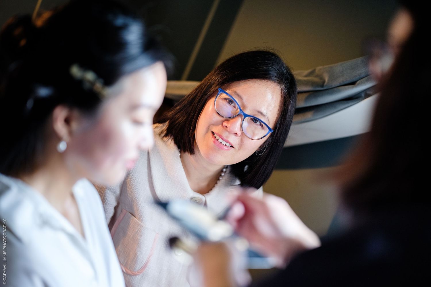 A smiling woman with blue glasses watches a makeup artist apply cosmetics to a bride during wedding preparations.