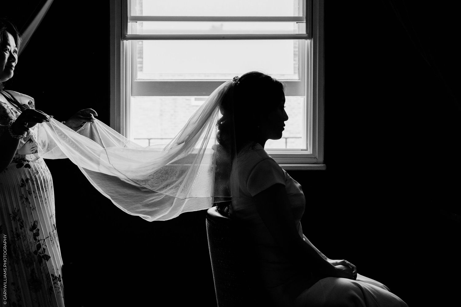 Silhouette of bride with flowing veil by window in black and white wedding photo.