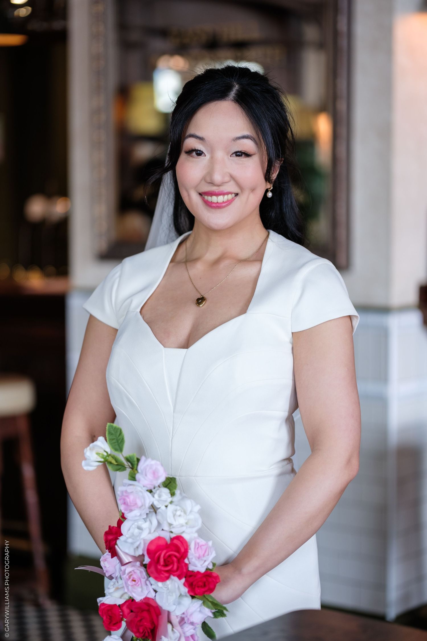 Smiling bride in white dress holds colorful bouquet of pink and red roses on her wedding day.