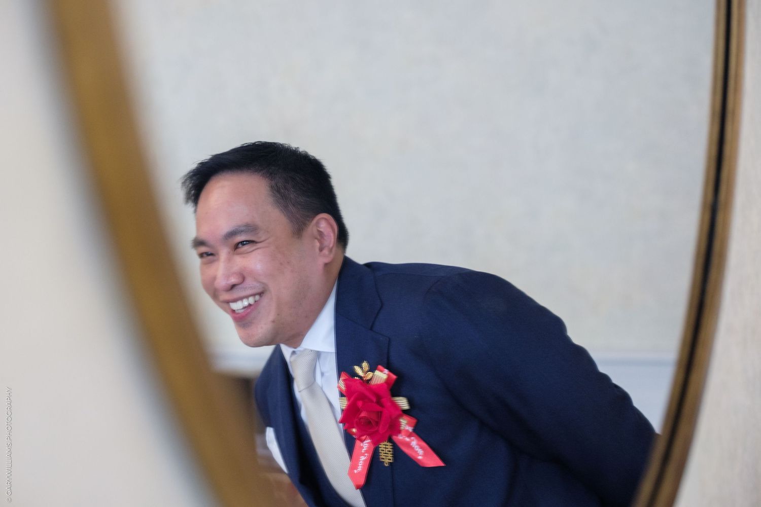 Smiling Asian groom in navy suit with red floral boutonniere laughing joyfully, reflected in elegant mirror.