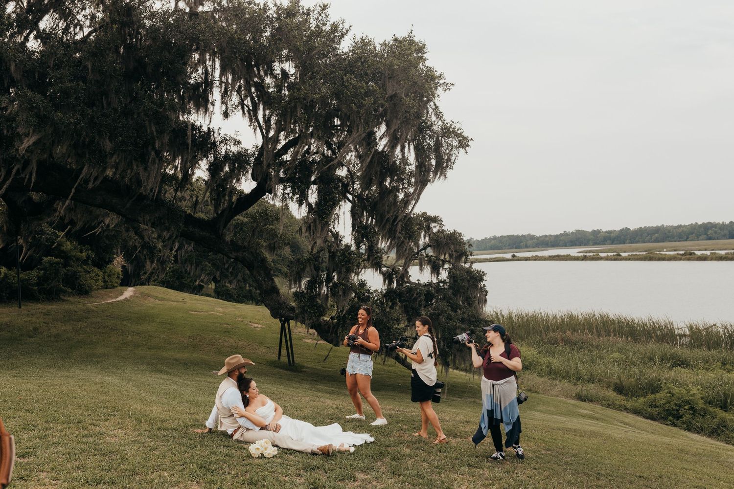 Group of friends enjoying a picnic on a grassy waterfront lawn under a large moss-draped tree.