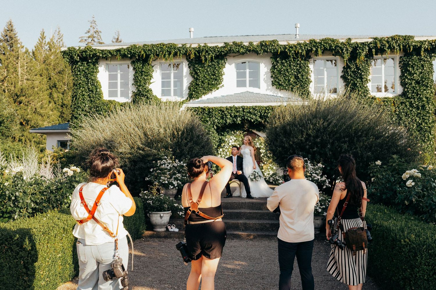 Wedding photographers capture a couple posing before an ivy-covered venue during an outdoor ceremony.