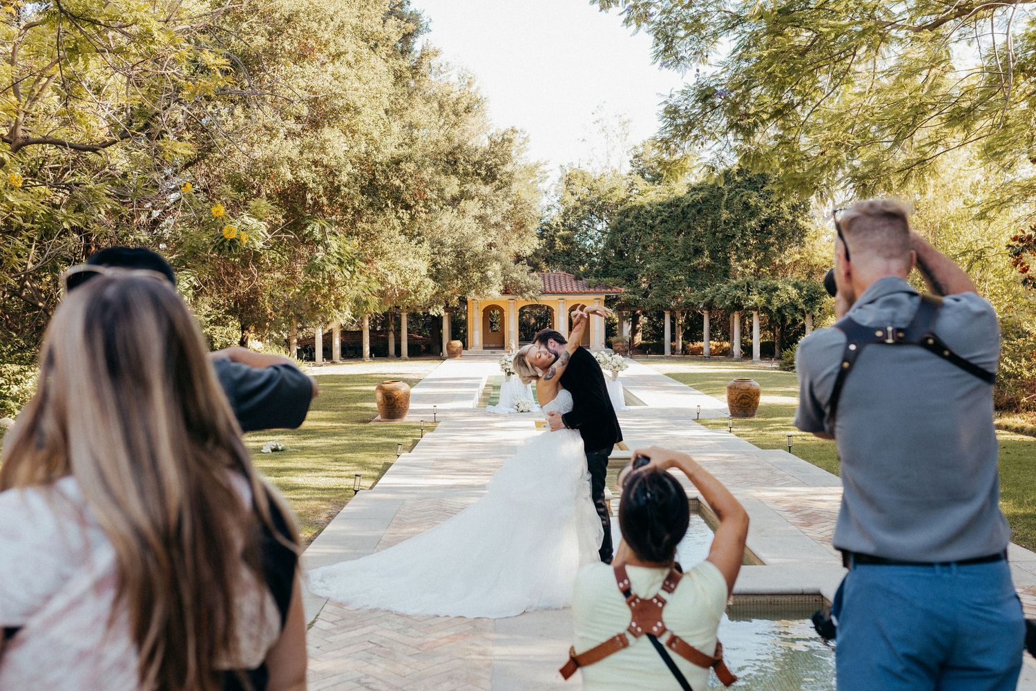 Wedding couple kissing outdoors while photographers capture the moment in a lush garden venue.