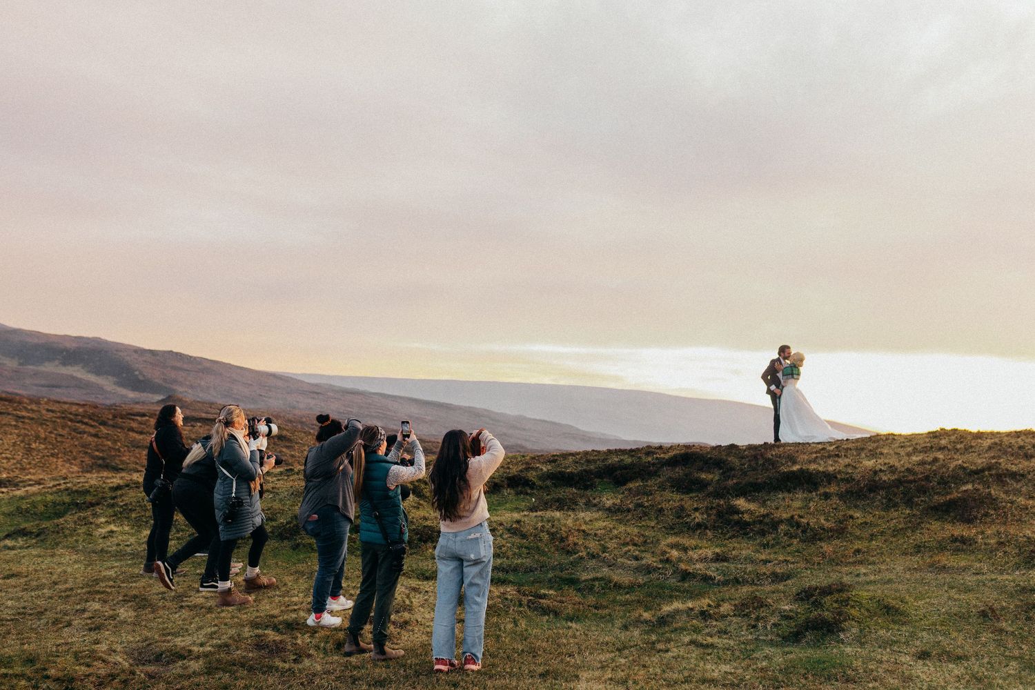 Group of people on a grassy hilltop at sunset with scenic mountain and ocean views in the background.
