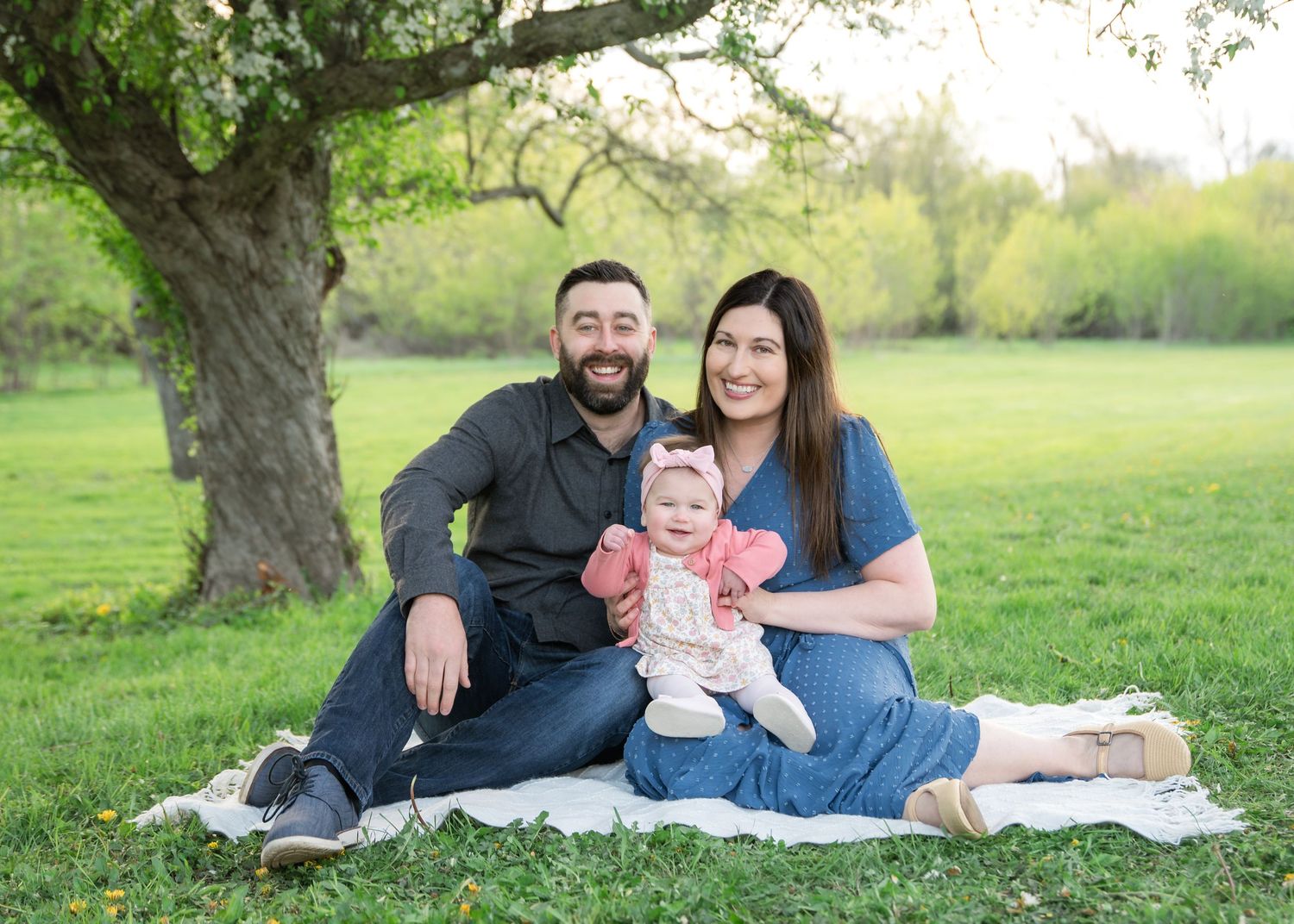 A happy family of three sits on a blanket under a blooming tree in a lush green park, smiling with their baby girl.