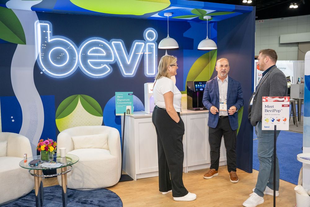 Three people networking at a Bevi beverage station booth display at a trade show or corporate event.
