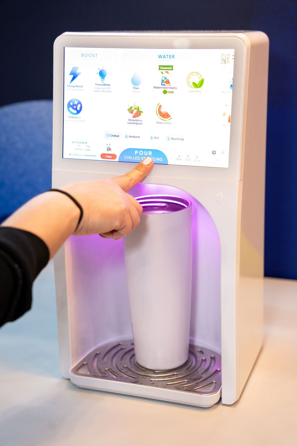 Person pressing button on modern touchscreen beverage dispenser with purple LED light and white cup.