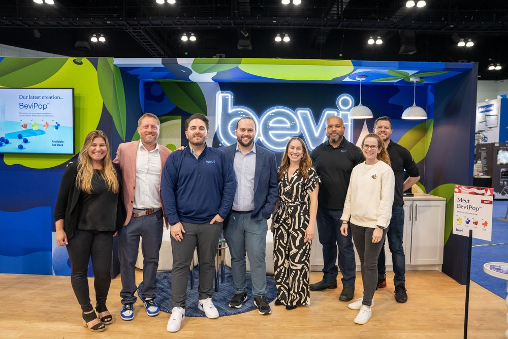 Group of seven professionals smiling together at a Bevi branded booth display at a trade show event.