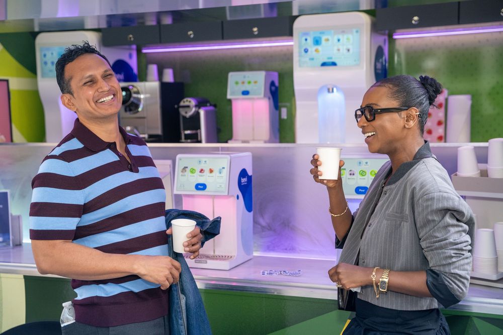 Two people smiling and holding coffee cups in a modern cafe with colorful machines in the background.