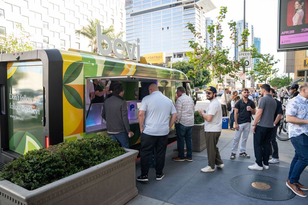 Crowd of people gathered around a colorful green and yellow food truck kiosk on a busy urban street.