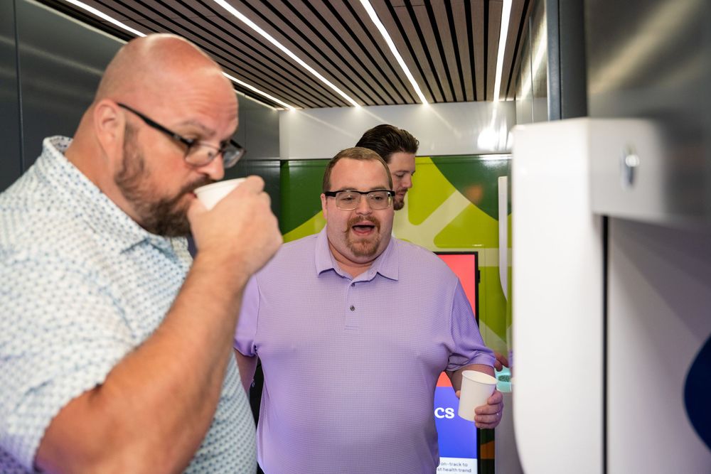 Two men sampling beverages at a modern self-serve drink dispenser machine indoors.