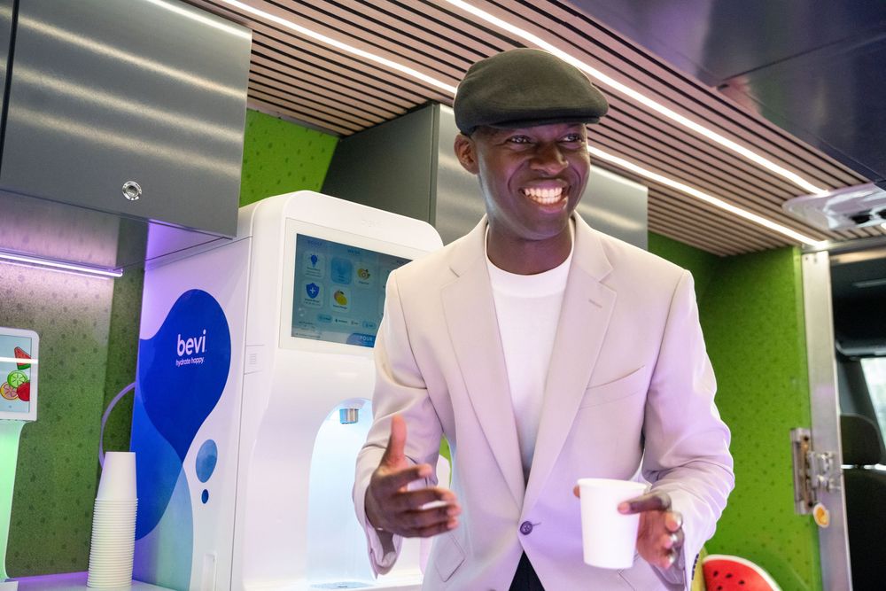 Man in beige blazer and cap smiling while holding a cup near a Bevi beverage dispenser machine.