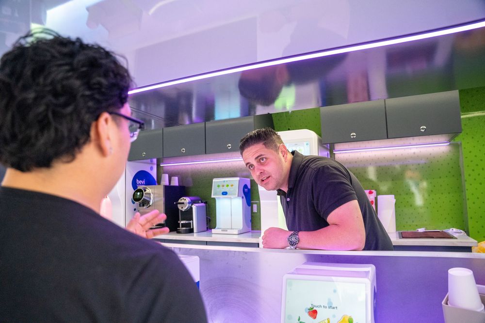 Two men conversing at a modern bar counter with green and purple decor in a contemporary cafe setting.