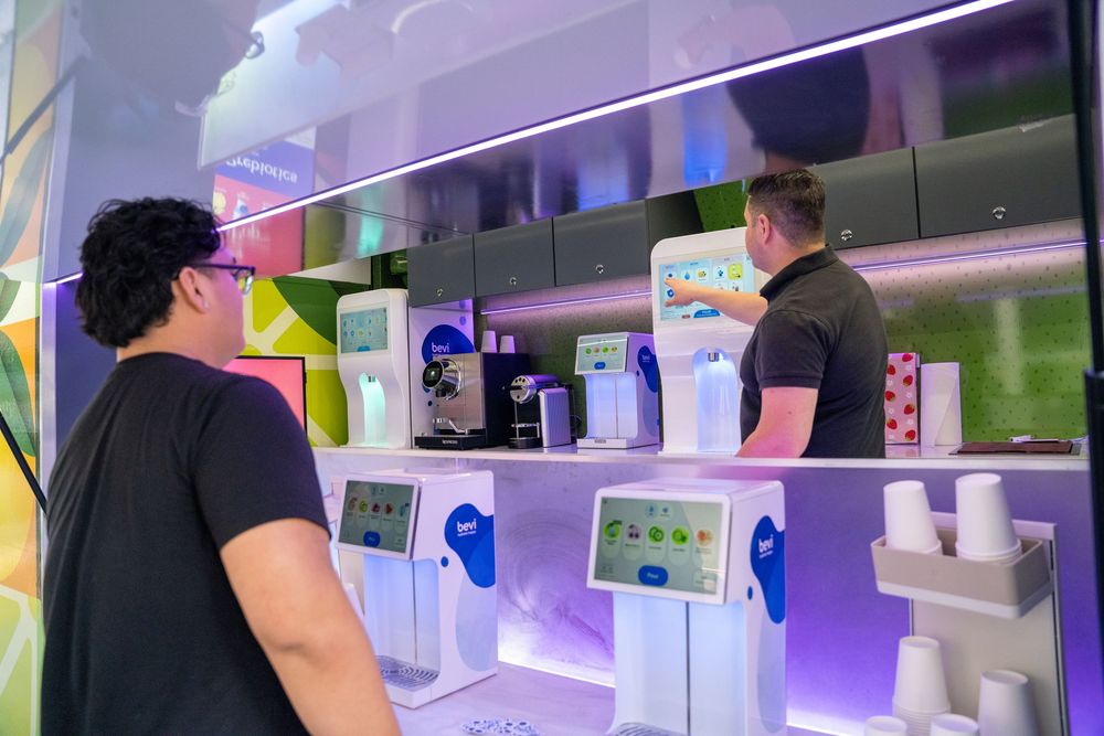 Two people ordering at a modern self-service kiosk counter in a colorful café or beverage shop.
