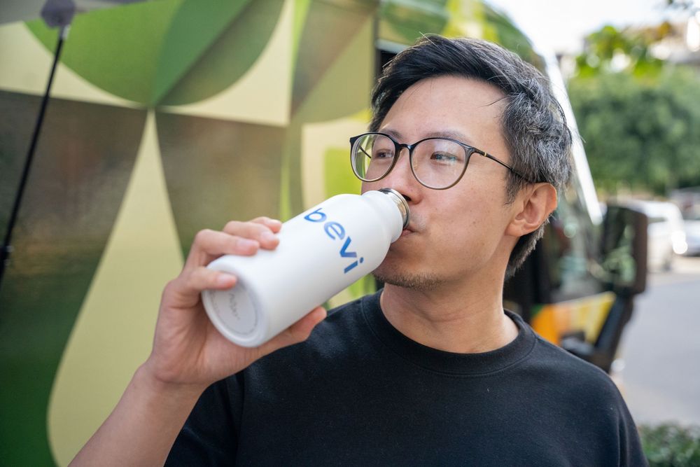 Asian man wearing glasses and black shirt drinking from a white branded cup outdoors.