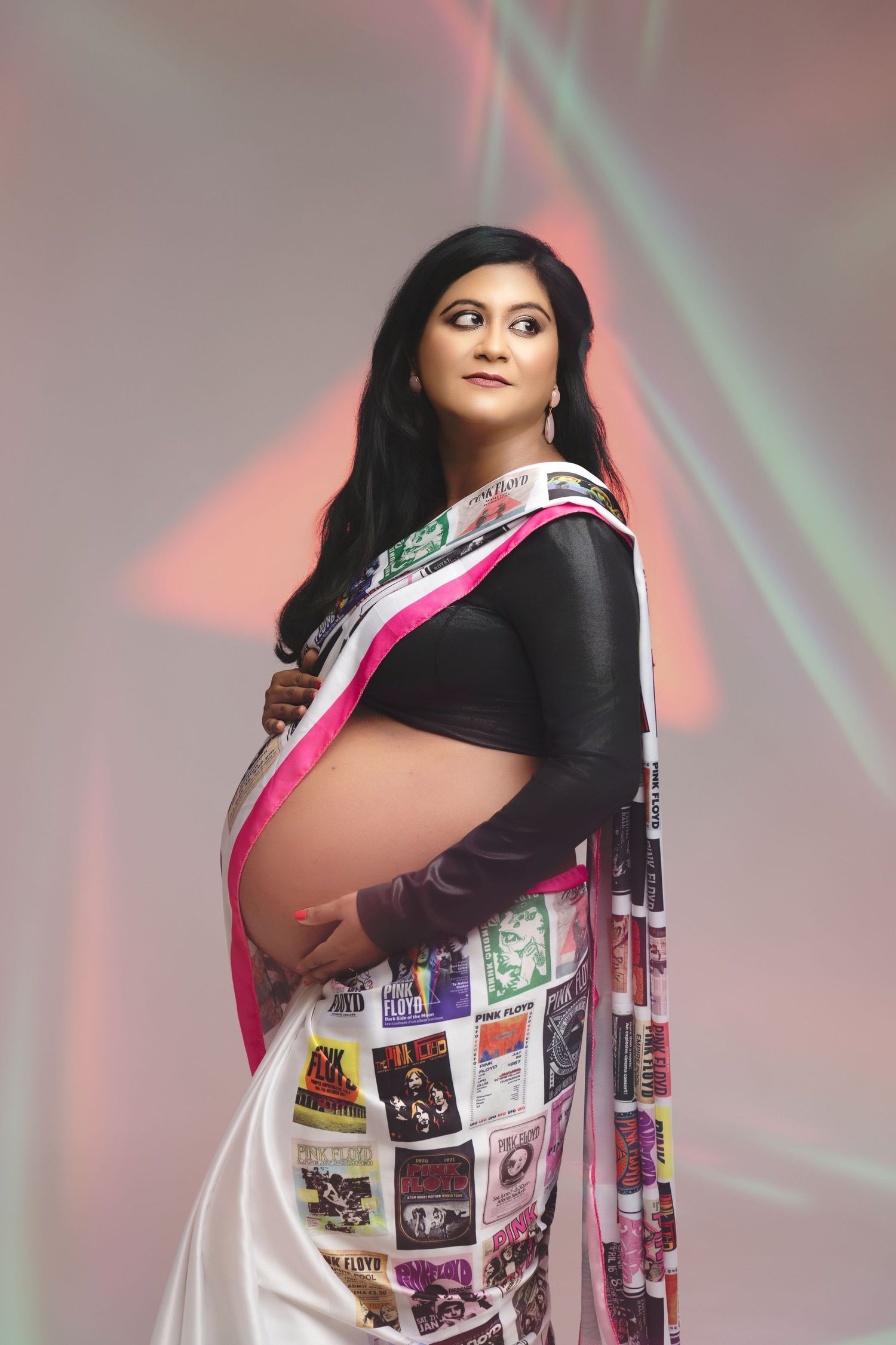 Pregnant woman in elegant white and pink saree with concert posters, posing against colorful light background.