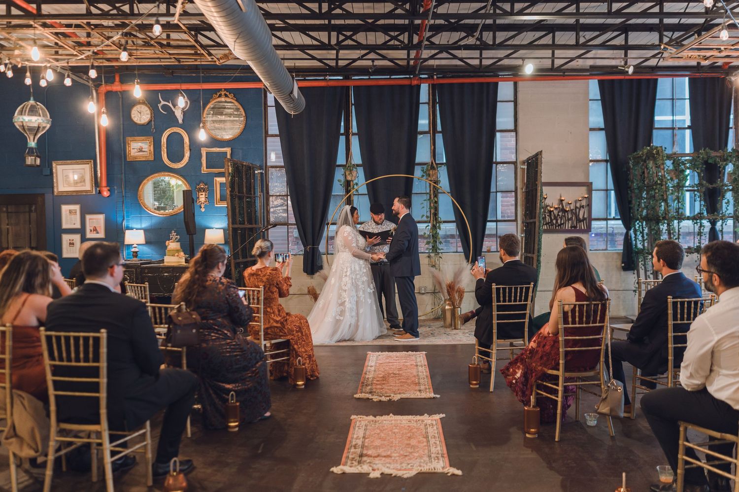 Couple exchanging vows at an indoor wedding ceremony with gold chairs, blue walls, and vintage gallery wall decor.