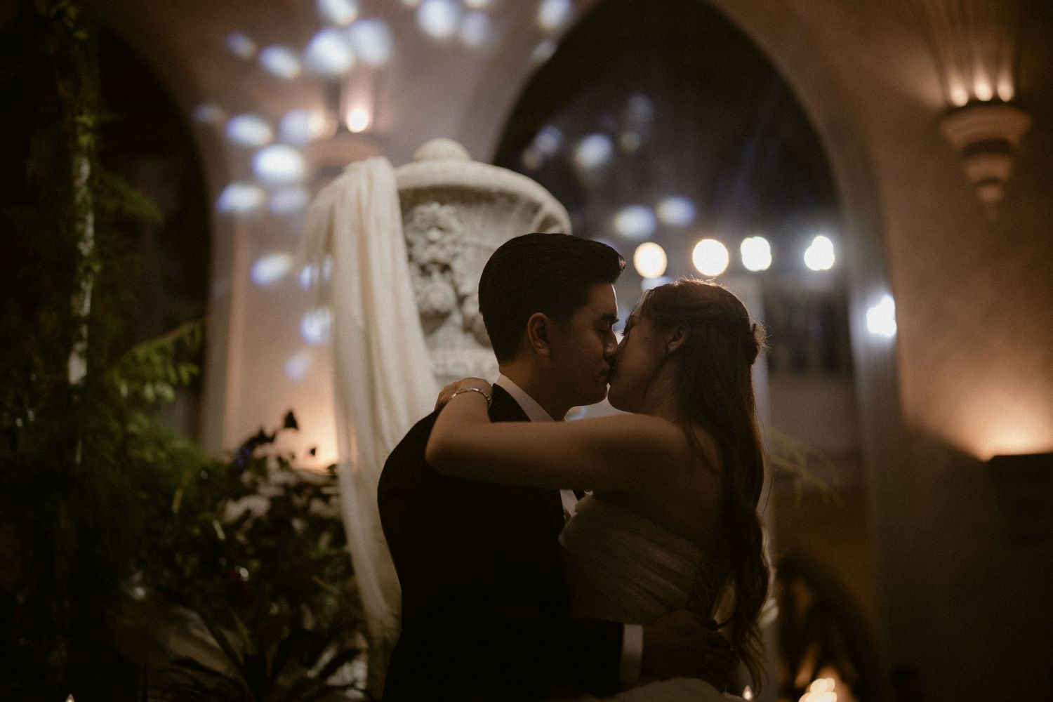 Bride and groom share a romantic kiss under soft bokeh lights in a warmly lit gothic venue.