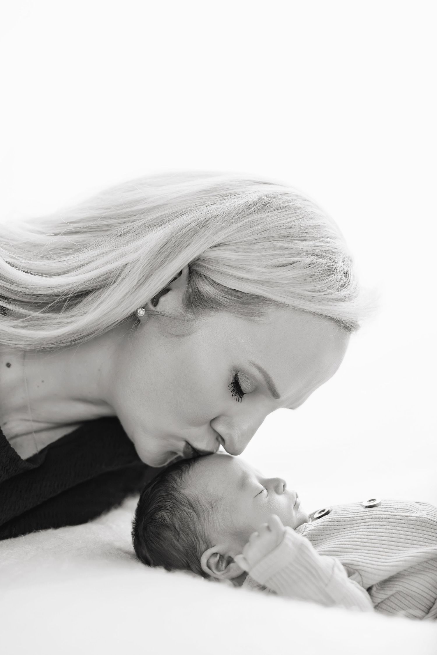 Black and white portrait of a mother kissing her sleeping newborn baby's forehead.
