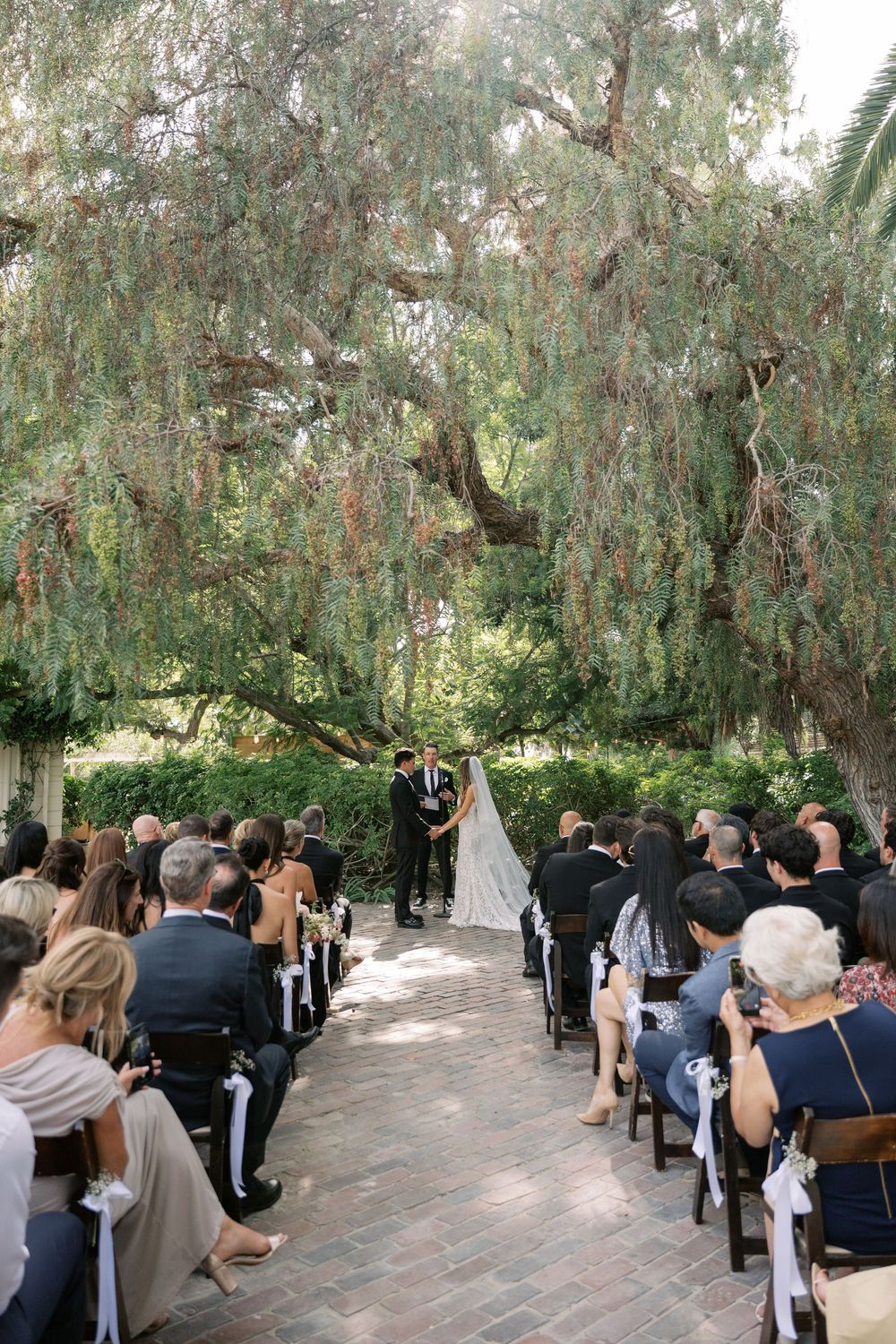 Outdoor garden wedding ceremony with guests seated along brick aisle beneath large leafy trees.