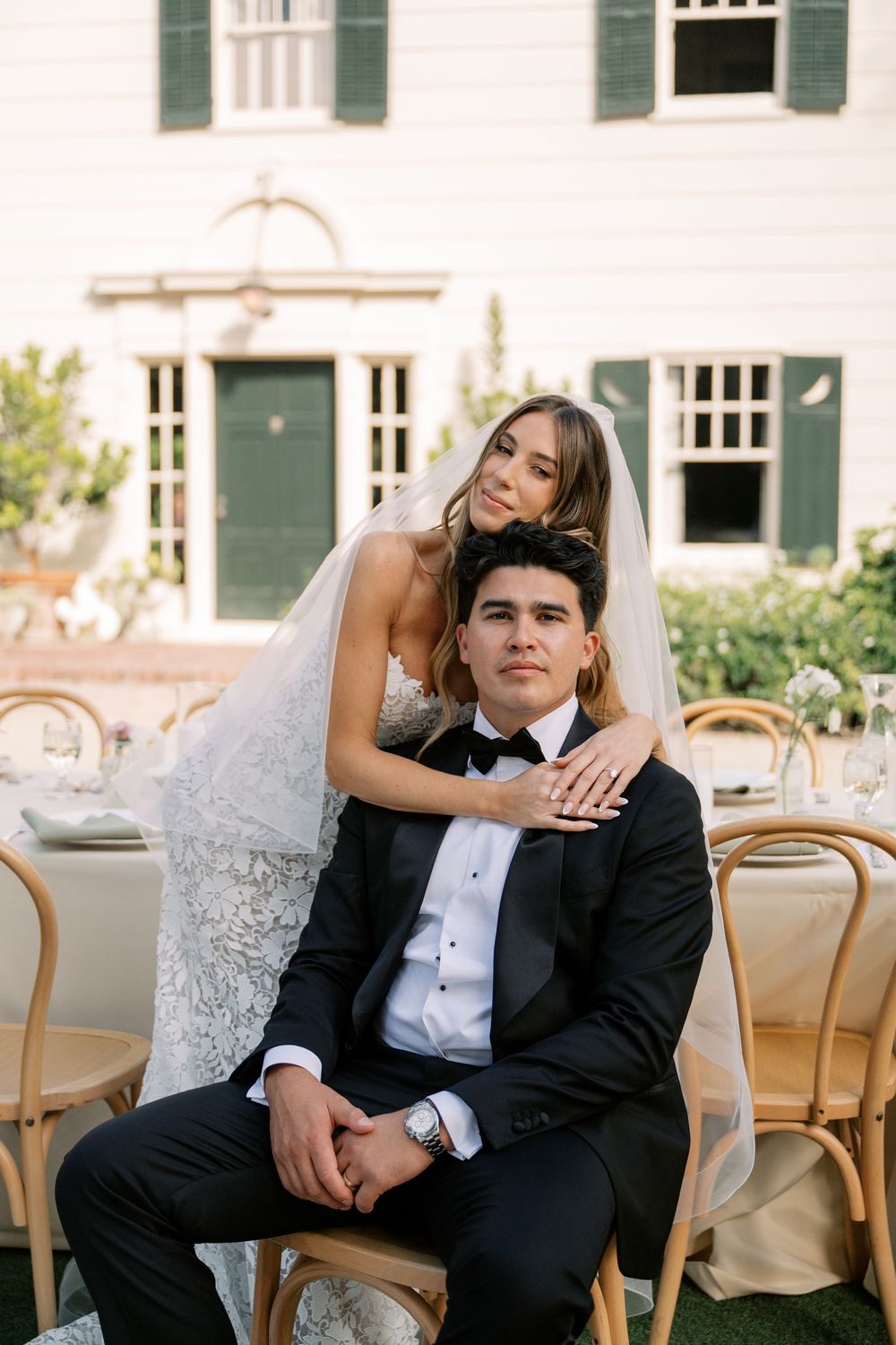 Bride in lace gown and veil embraces groom in black tuxedo seated at outdoor wedding reception table.