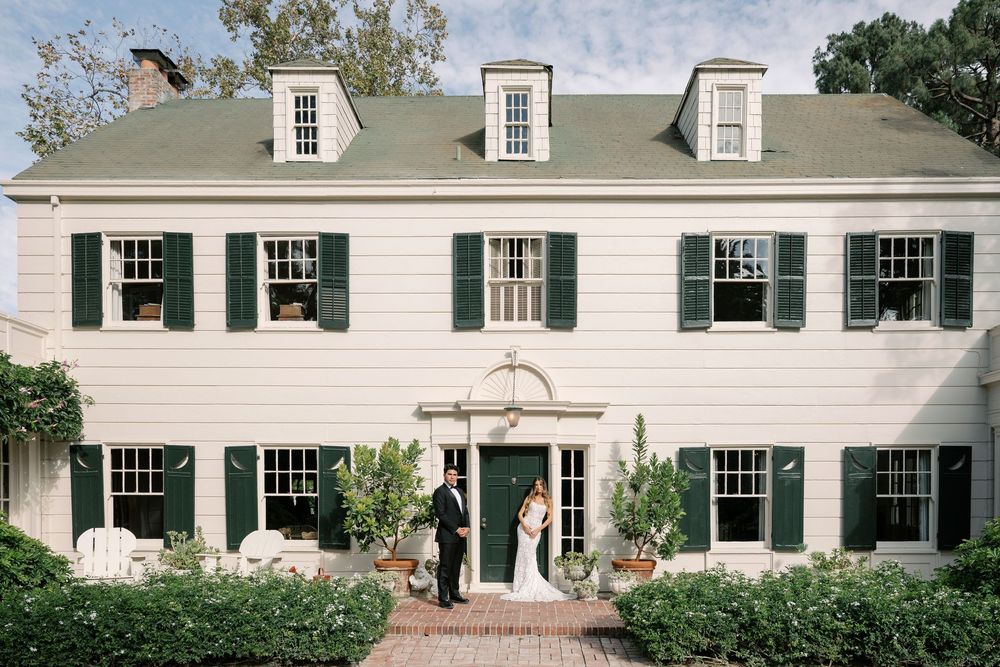 Bride and groom stand before an elegant white colonial home with green shutters.