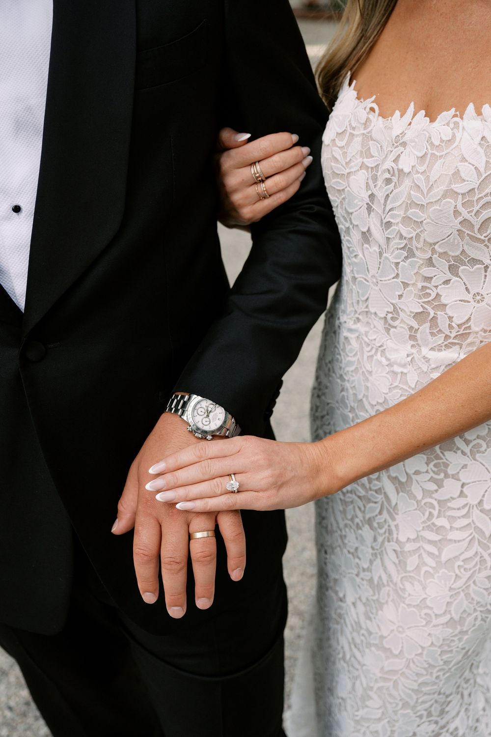 Bride and groom displaying wedding rings, bride in lace gown, groom in black tuxedo, close-up of hands.