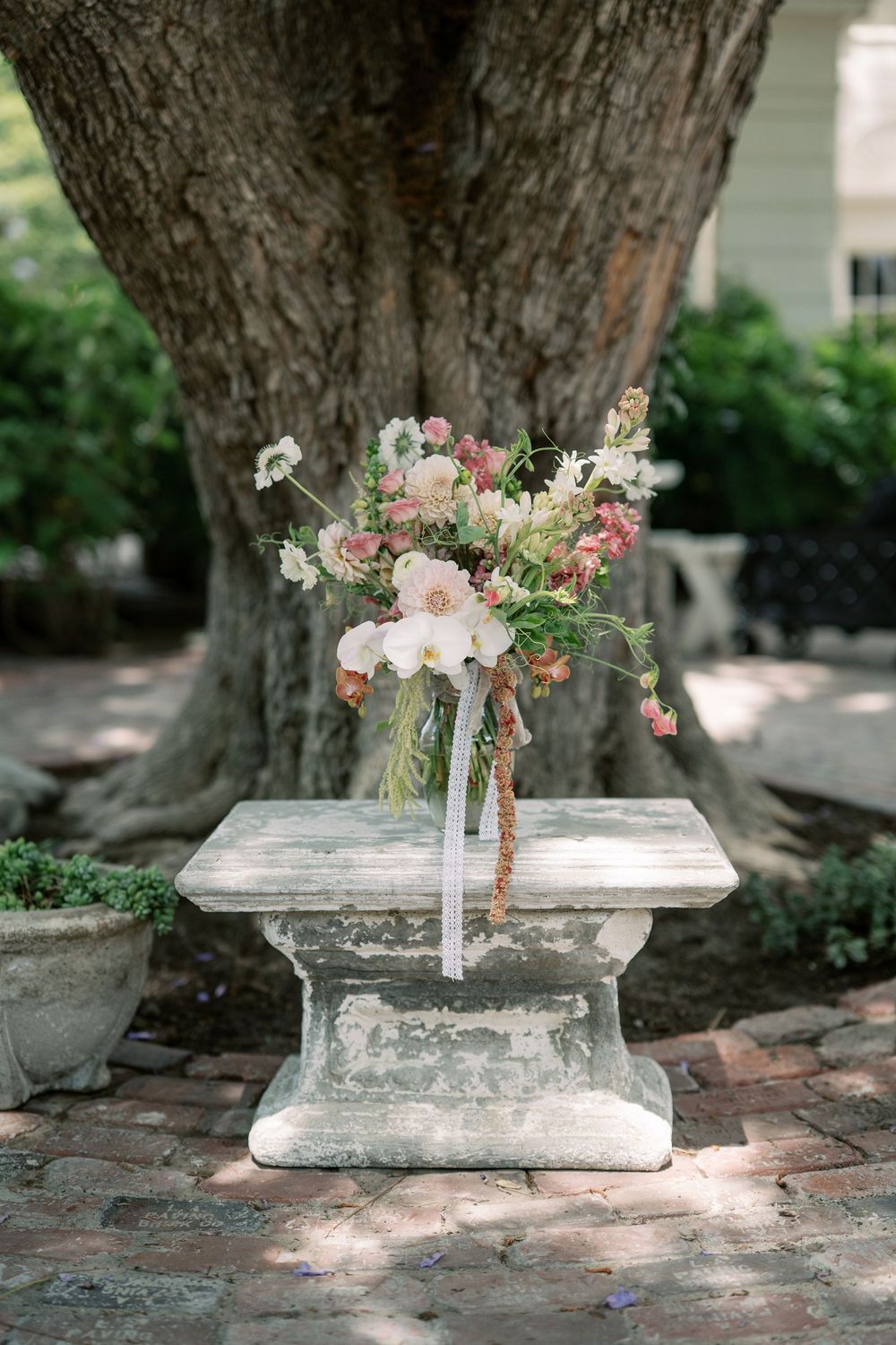 Romantic pink and white floral bouquet rests on stone pedestal beneath large tree at garden wedding.