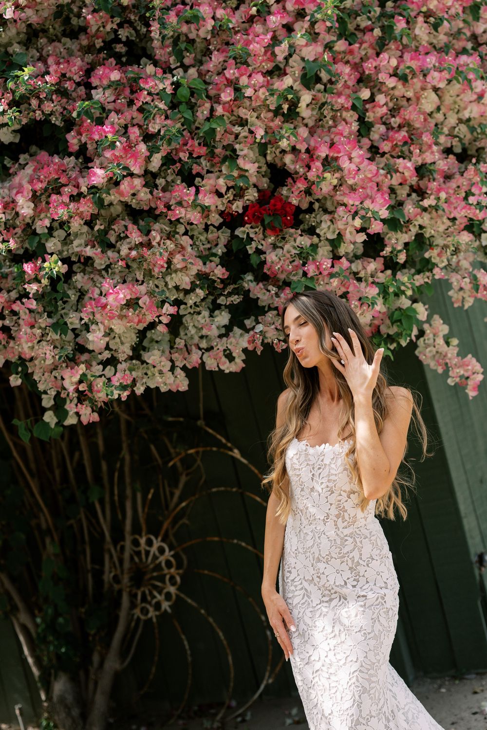 Bride in lace mermaid gown posing before vibrant pink bougainvillea blooms, long flowing hair, elegant outdoor bridal portrait.