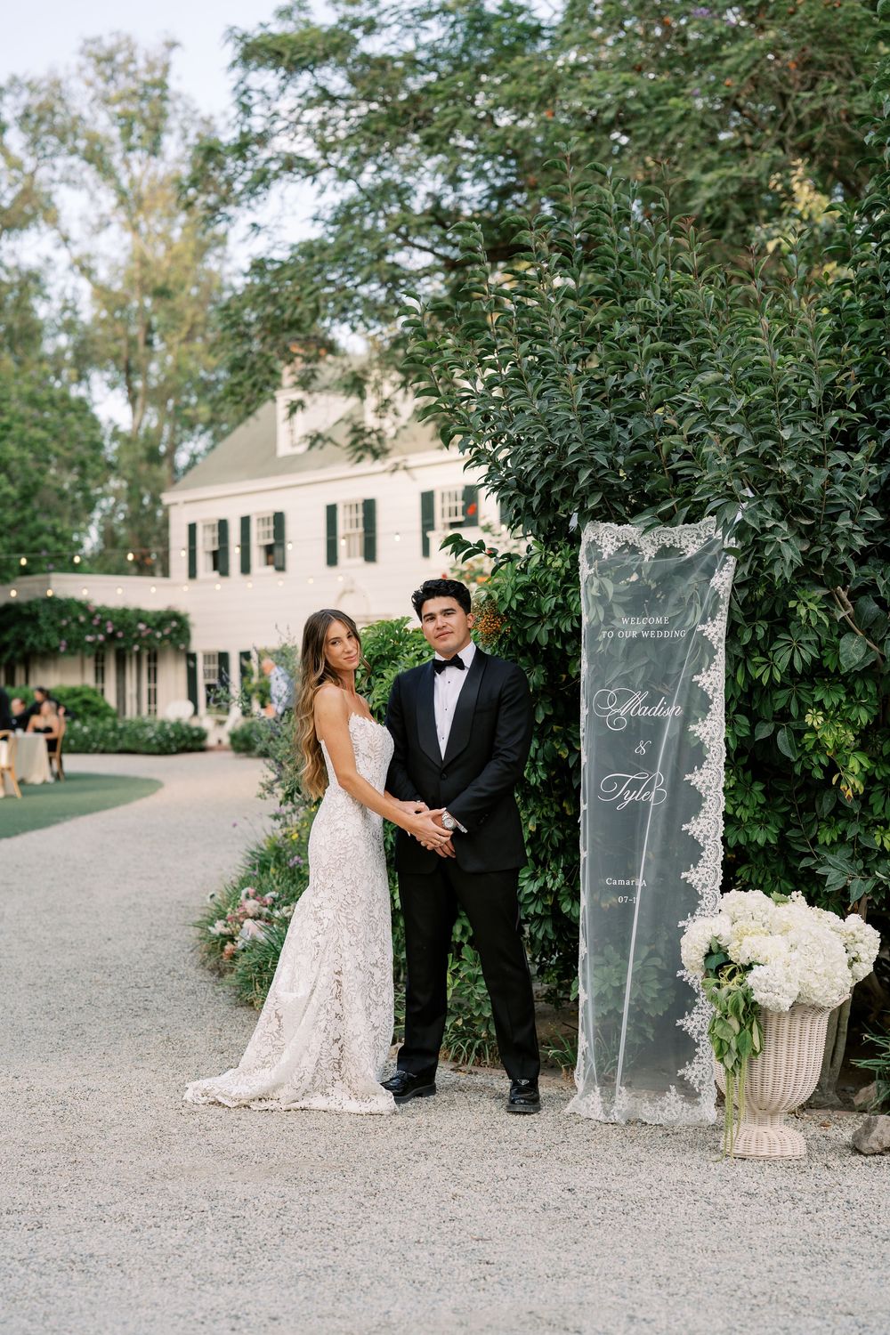 Bride in lace gown and groom in tuxedo pose beside elegant acrylic wedding welcome sign outside white venue.