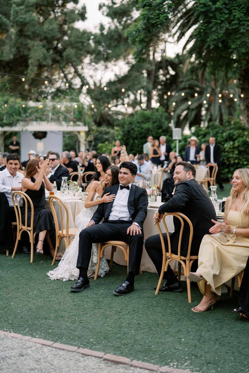 Bride and groom laugh with guests at outdoor wedding reception dinner under string lights at dusk.