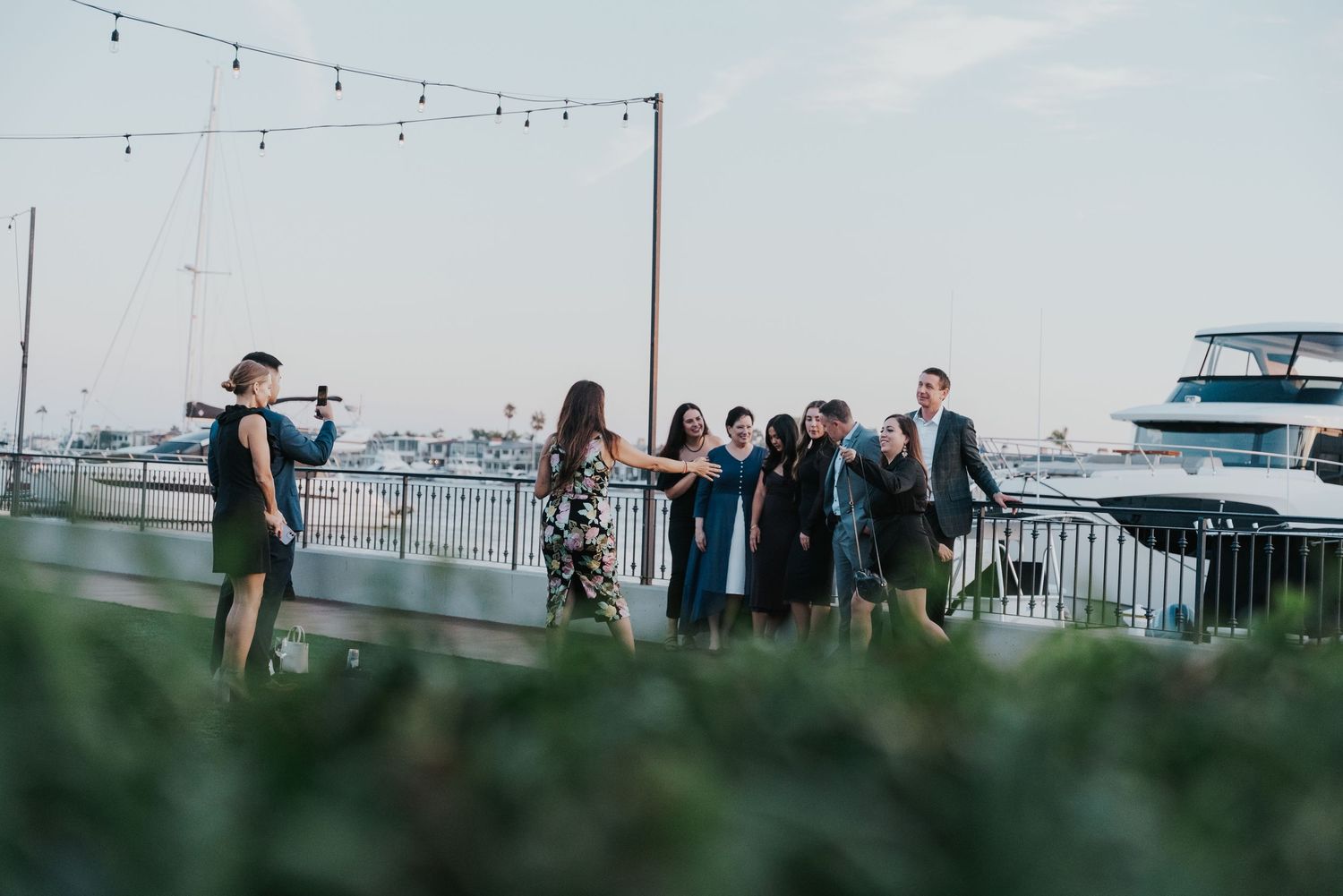 Group of elegantly dressed guests socializing on a waterfront terrace at dusk with boats and string lights in background.