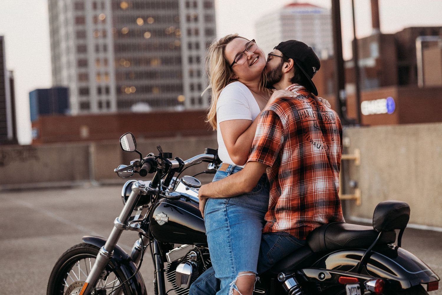 A couple shares a romantic moment on a motorcycle on a rooftop parking lot with a city skyline in the background.