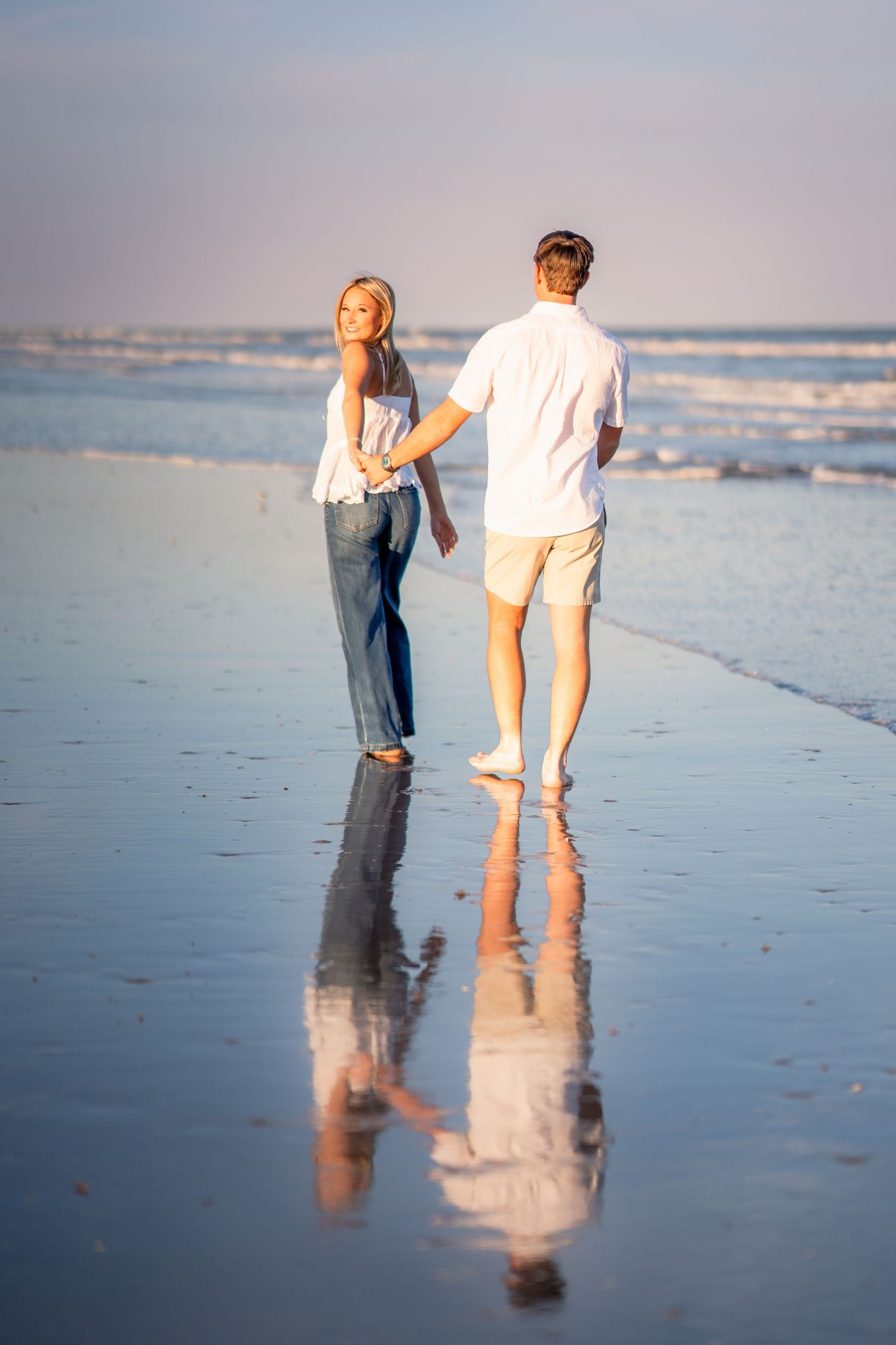 Couple holding hands walking barefoot on a reflective wet beach at sunset, woman in jeans glancing back over shoulder.