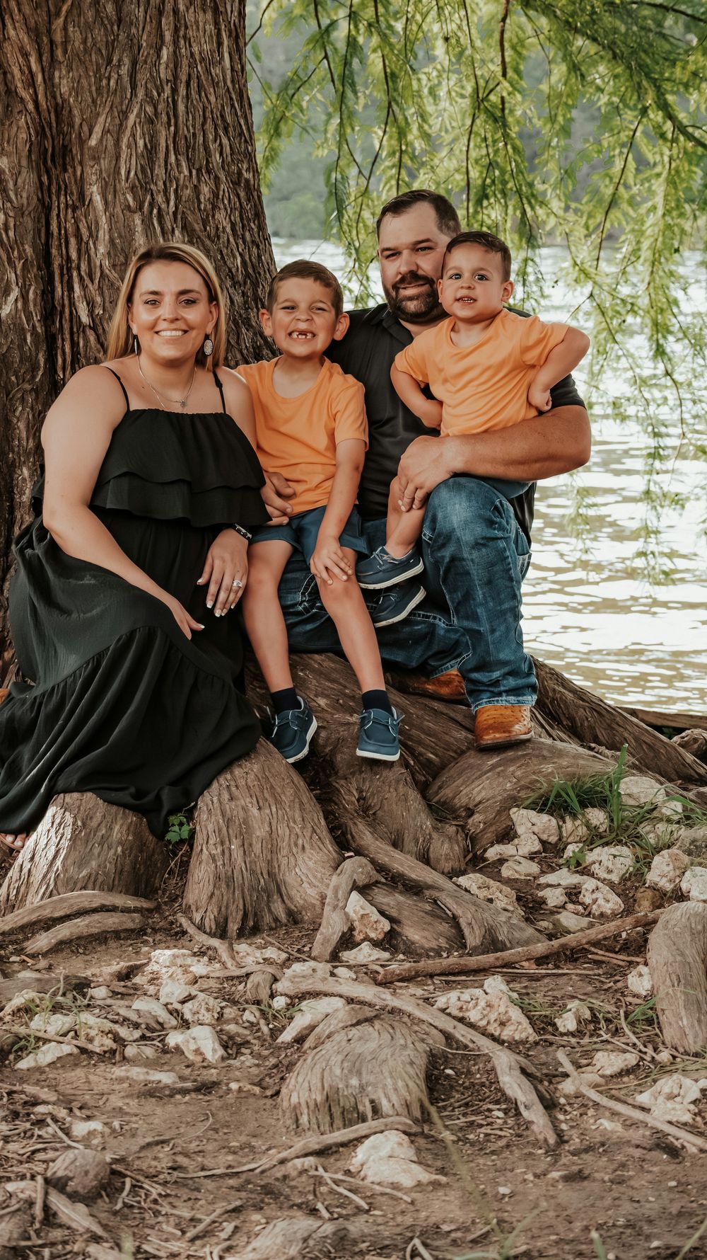 A happy family of four poses together under a large tree near a river for a portrait.