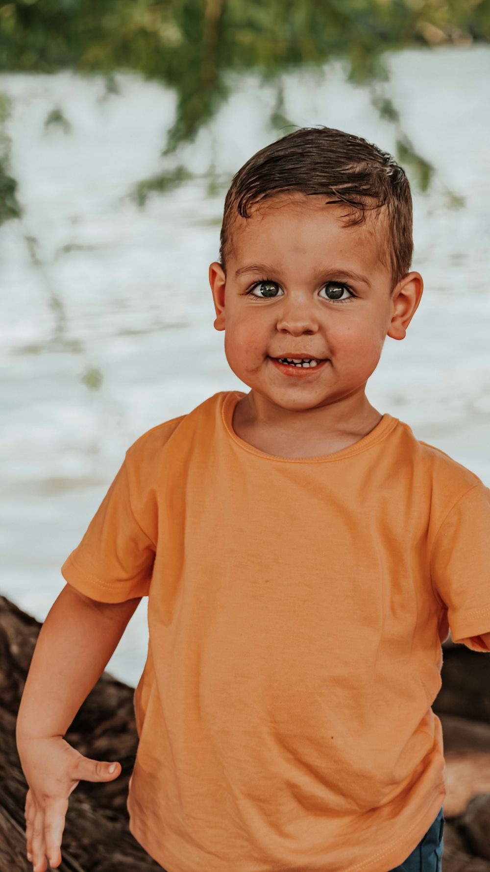 Adorable toddler boy in an orange t-shirt smiling at the camera near a sunlit water background.