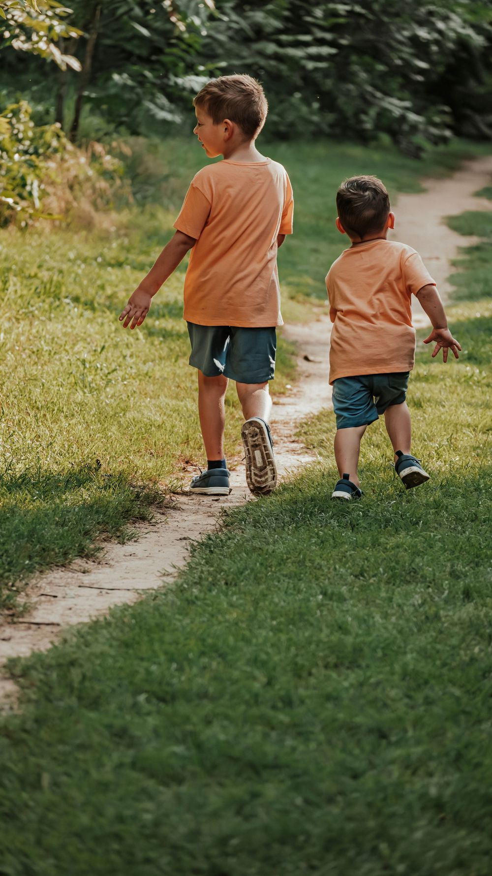 Two young boys in matching orange shirts and blue shorts walk along a winding garden path surrounded by lush green grass.