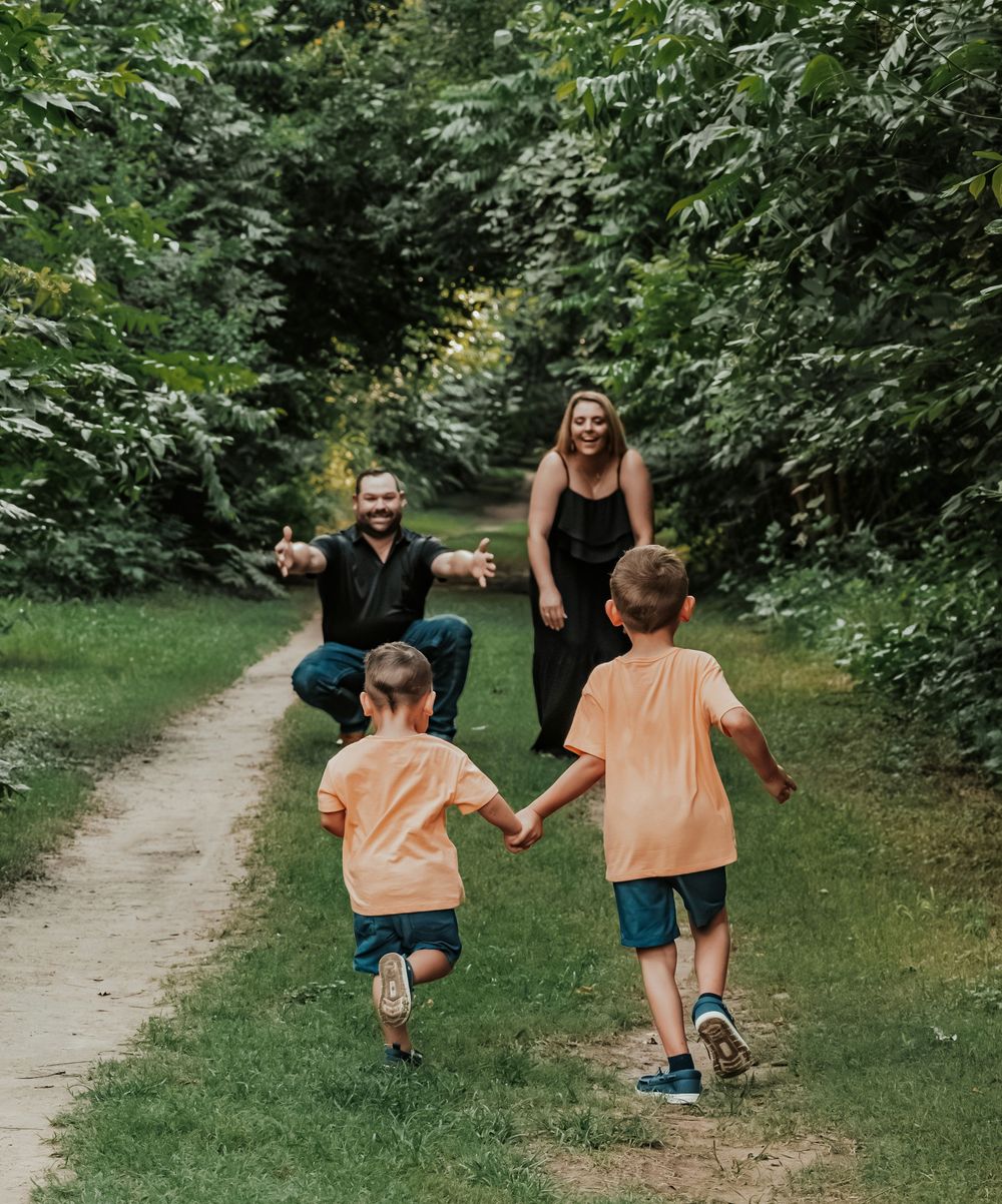 Two children in orange shirts run toward their smiling parents on a lush green park path.