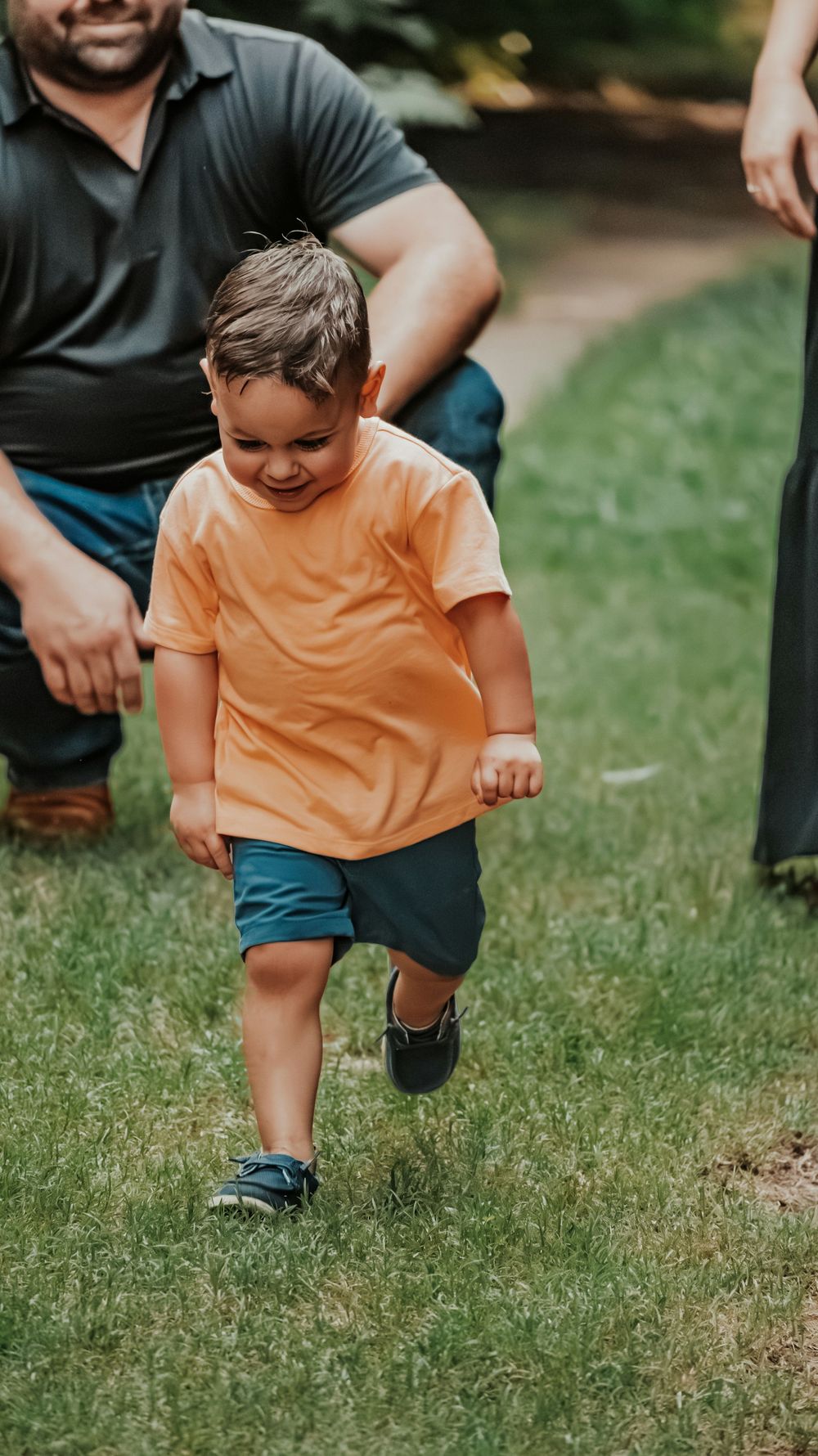 A happy toddler in an orange shirt and blue shorts runs on green grass during a family outdoor photo session.